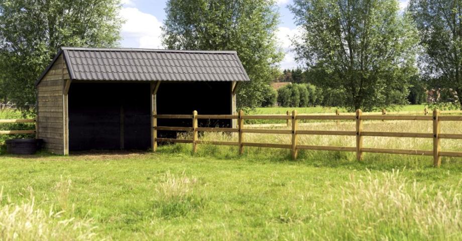 Un double abri à chevaux en bois avec des tuiles noires relié à des clôtures en bois à 3 rails